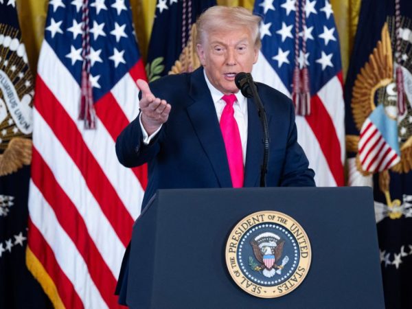US President Donald Trump speaking at a podium with American flags and presidential seals in the background.