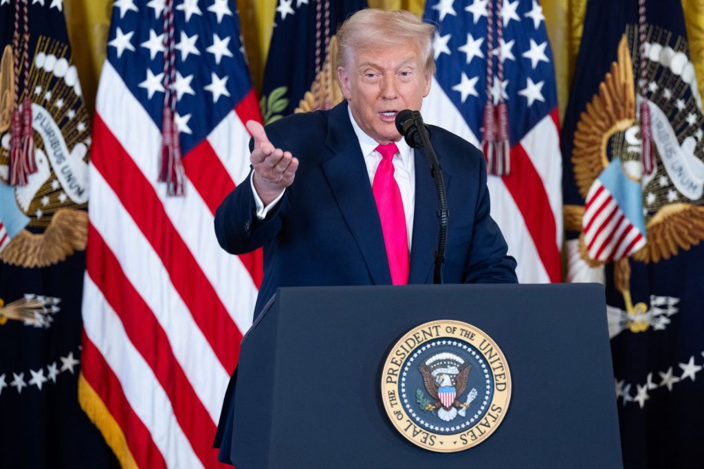 US President Donald Trump speaking at a podium with American flags and presidential seals in the background.